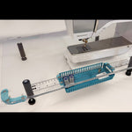 A sewing machine accessory setup on a white surface, featuring a clear acrylic guide rail with black adjustment knobs, a teal plastic basket, and a red-handled tool, with a Bernina sewing machine visible in the background.