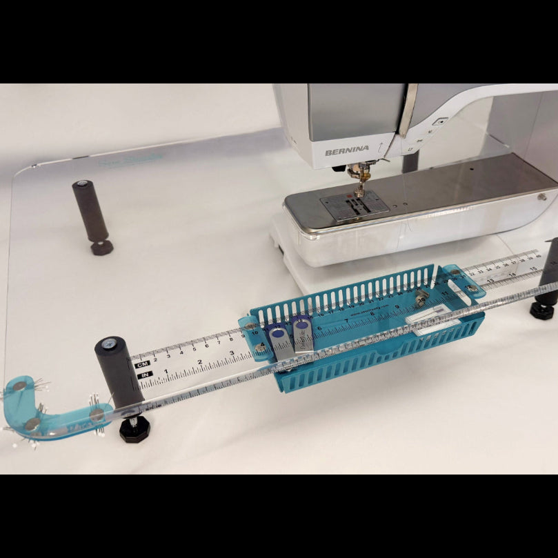 A sewing machine accessory setup on a white surface, featuring a clear acrylic guide rail with black adjustment knobs, a teal plastic basket, and a red-handled tool, with a Bernina sewing machine visible in the background.
