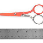 Red-handled scissors with silver metal blades and handles, shown above a metal ruler on a white background.