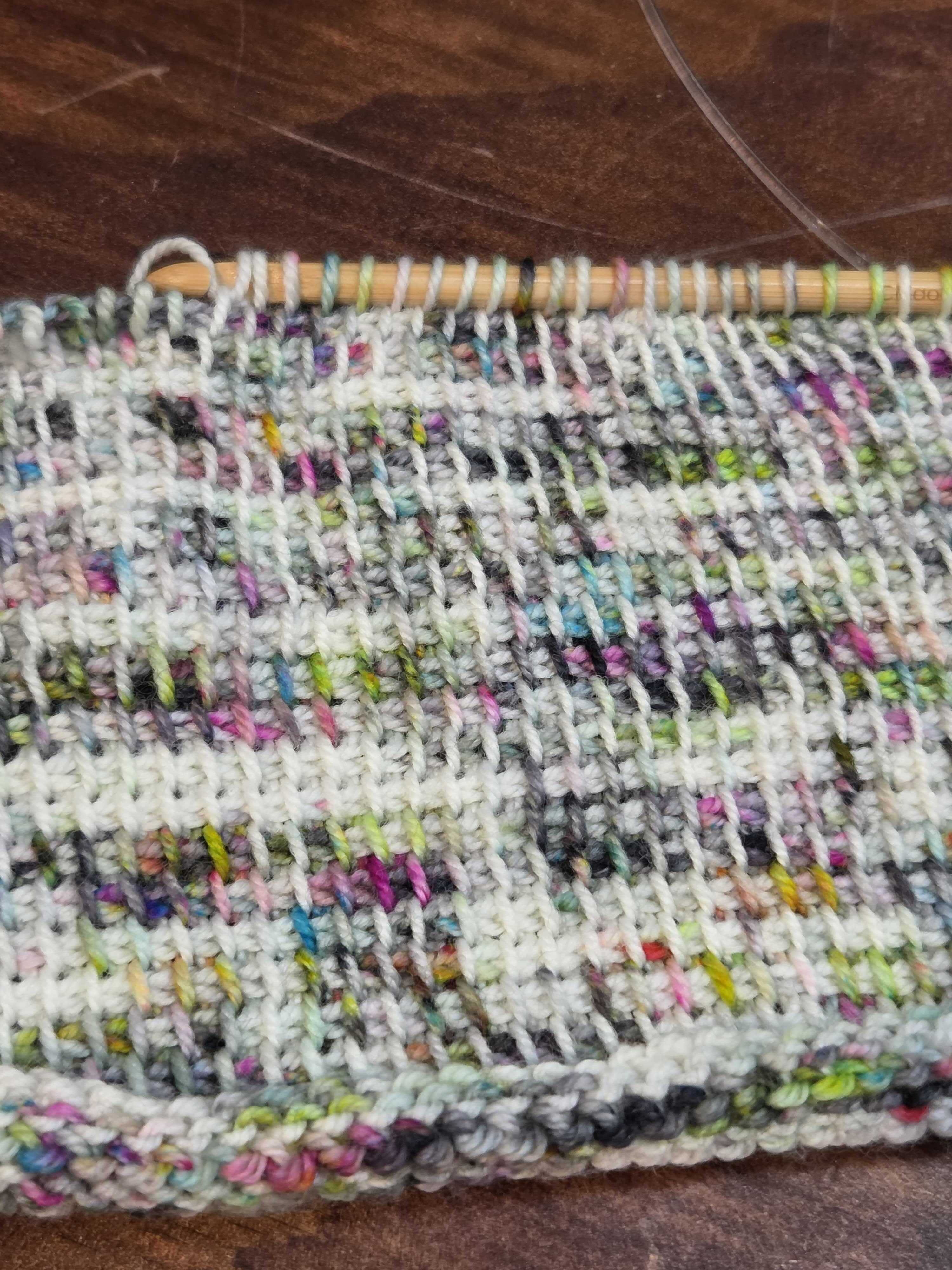 Tunisian crochet swatch in multicolored yarn, shown mid‑row with the hook holding active stitches at the top.