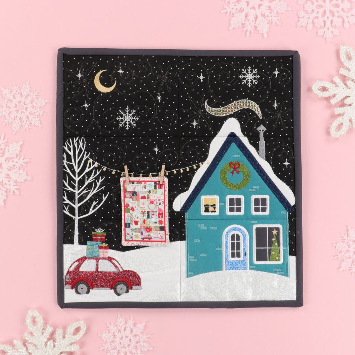 Quilted scene with a house, car, and tree on a pink background with snowflakes.