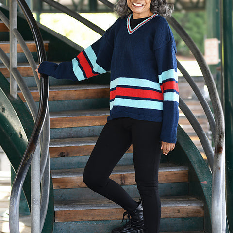 A smiling woman with gray hair and glasses stands on outdoor wooden stairs wearing a navy blue V-neck sweater with red, light blue, and white stripes, black leggings, and black boots.