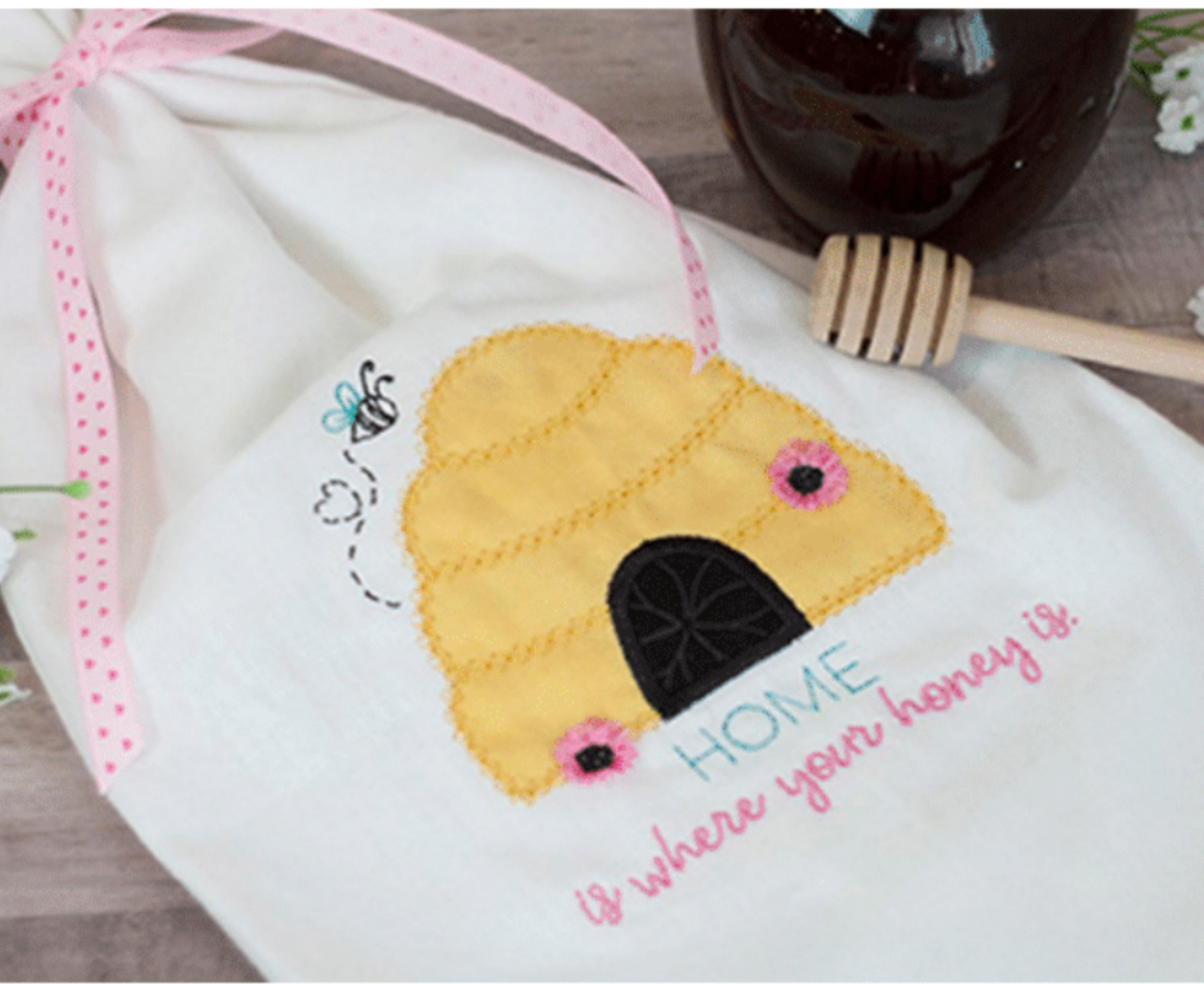 Embroidered bee hive design on a white fabric with honey dipper and jar in the background.
