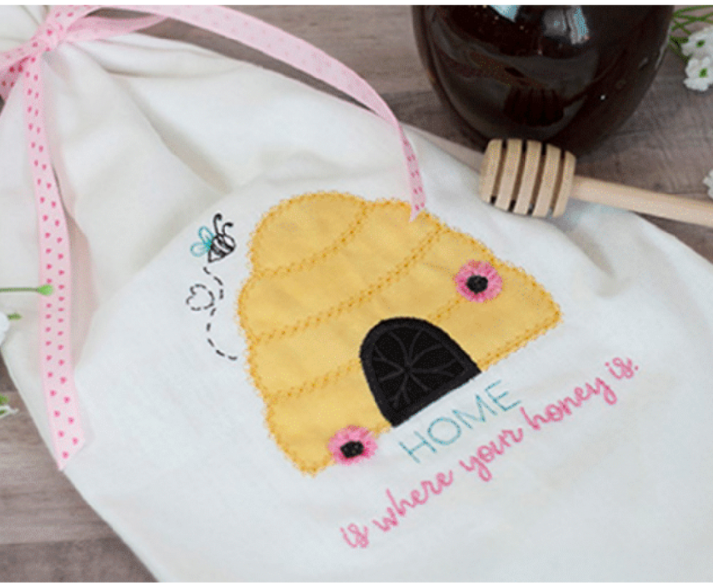 Embroidered bee hive design on a white fabric with honey dipper and jar in the background.