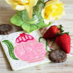 Coaster with pink teapot design, strawberries, and cookies on a wooden surface with flowers in the background.