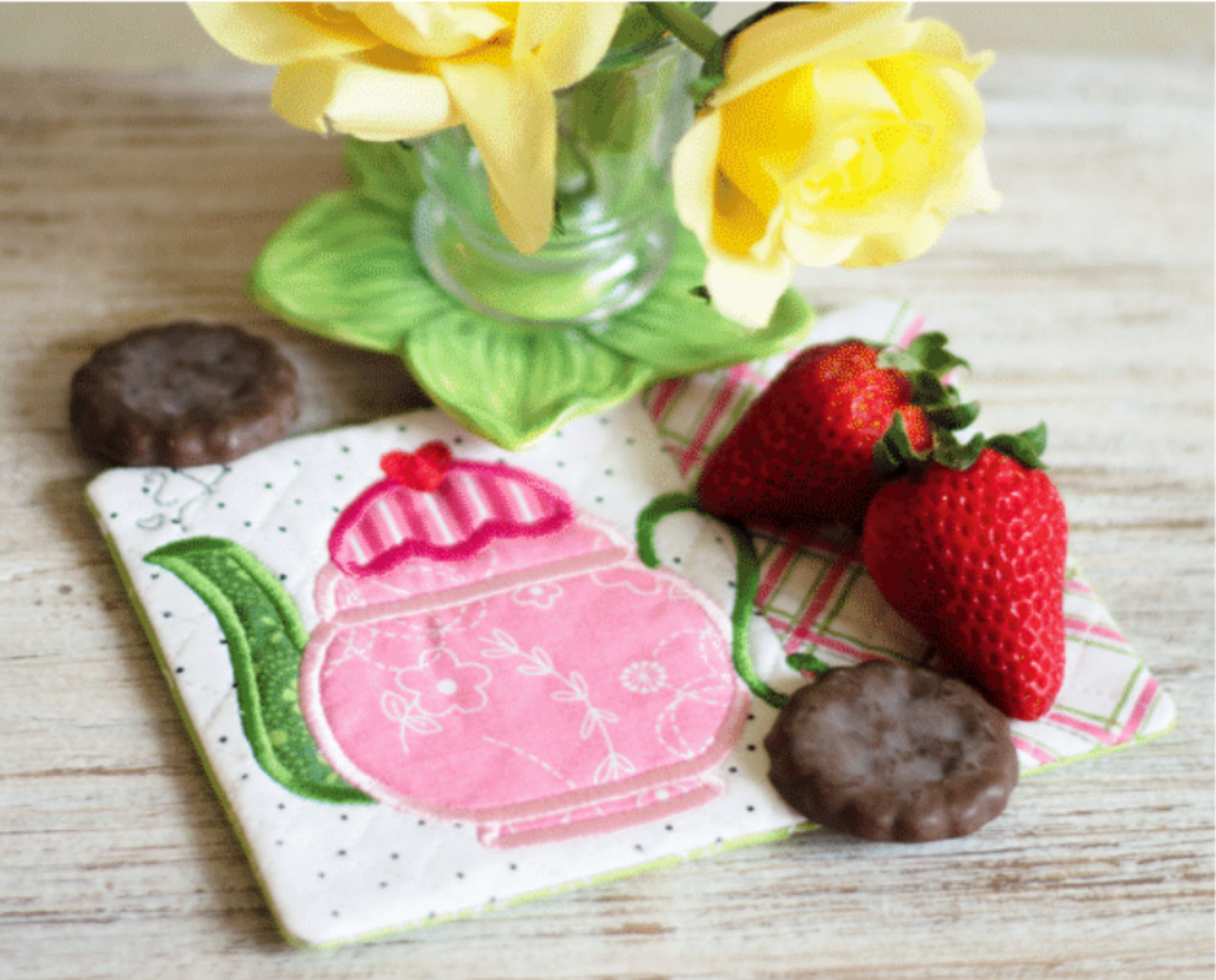 Coaster with pink teapot design, strawberries, and cookies on a wooden surface with flowers in the background.