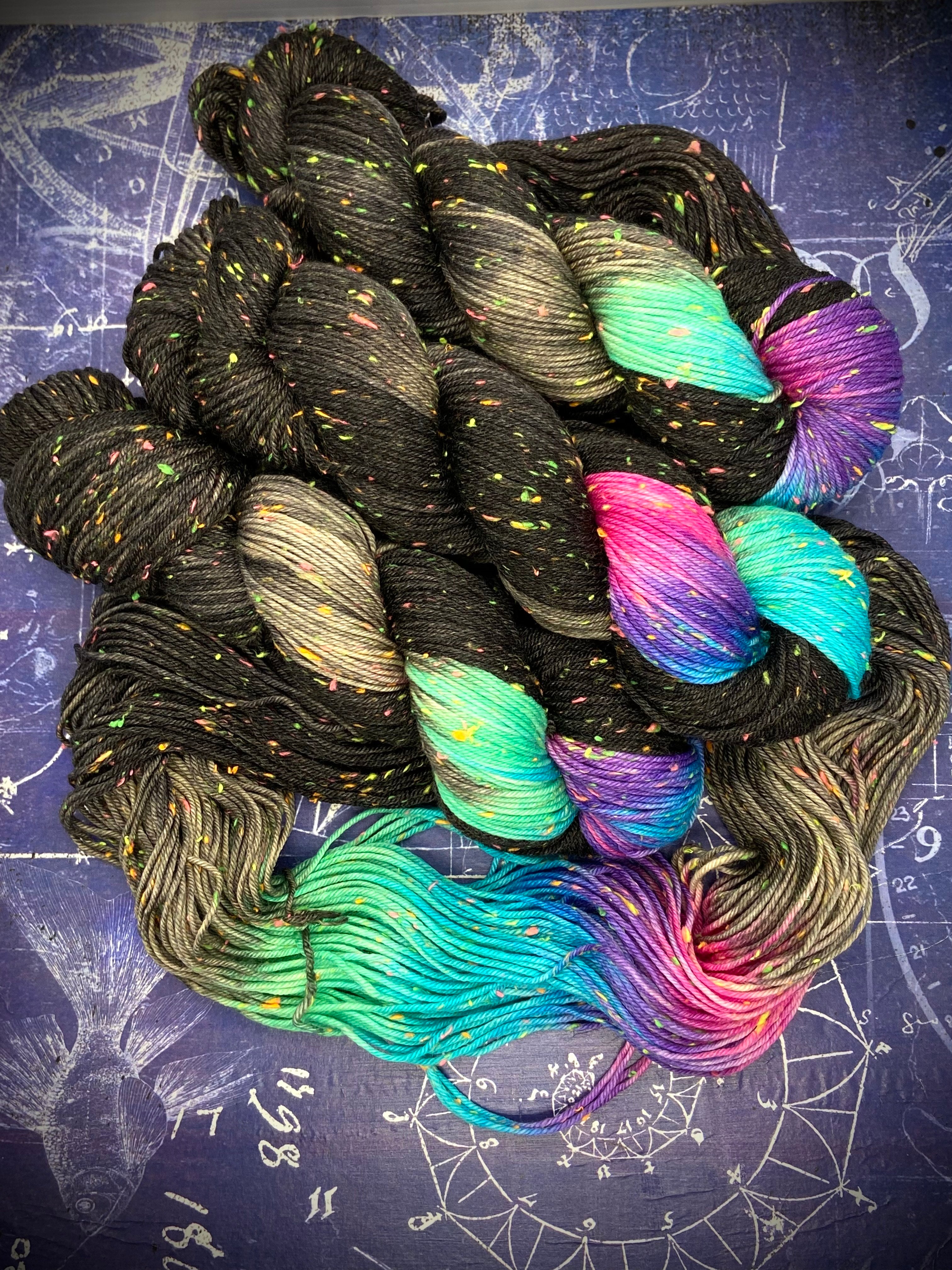 Black hand‑dyed yarn with multicolored neon sections arranged on a dark constellation‑themed background.