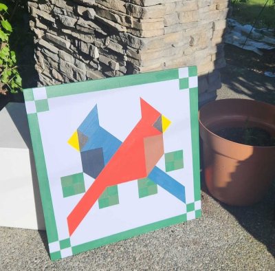 Painted barn quilt featuring two geometric birds in red and blue perched on green squares with a decorative border.