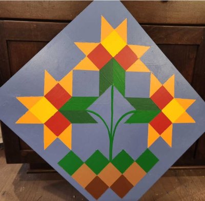 Painted barn quilt featuring three geometric floral motifs in yellow, orange, red, and green on a blue background.