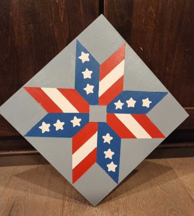 Painted barn quilt featuring a red, white, and blue geometric star design with striped and starred points.