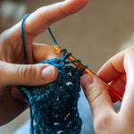 Hands crocheting with a hook and blue‑green yarn in a close‑up view.