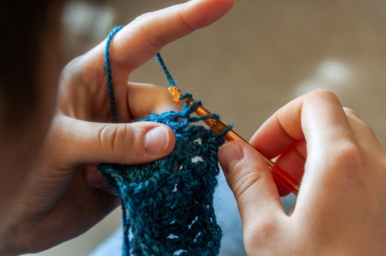 Hands crocheting with a hook and blue‑green yarn in a close‑up view.