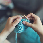 Hands knitting with wooden needles and light blue yarn in a close‑up view.