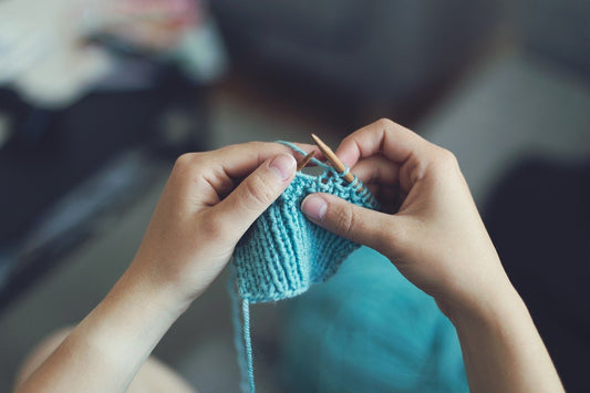Hands knitting with wooden needles and light blue yarn in a close‑up view.