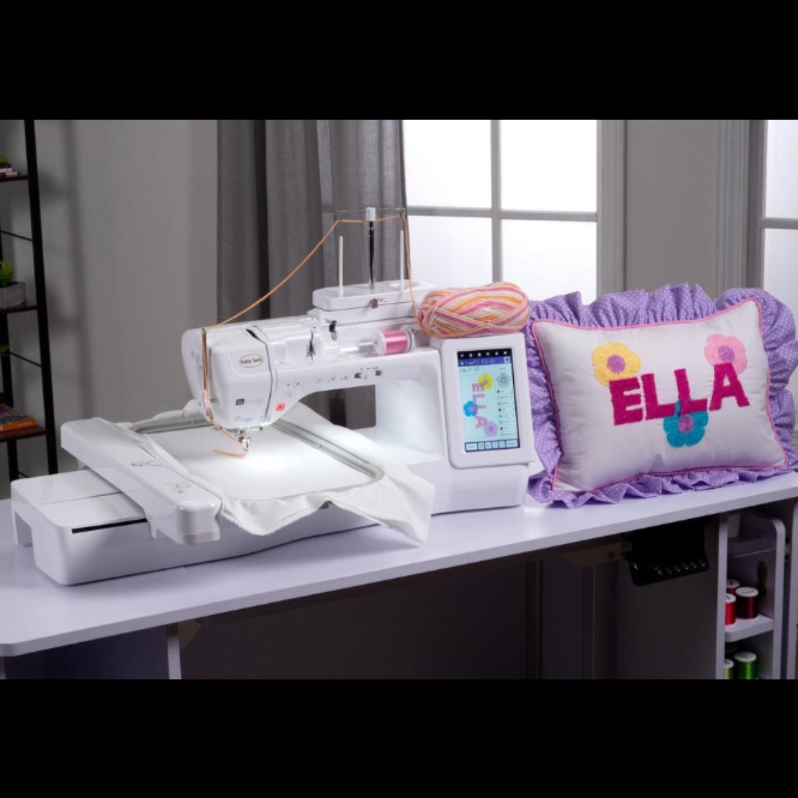 A white embroidery machine sits on a white table in a bright room, with a white pillow featuring a purple ruffled border and colorful embroidered text reading 'ELLA' beside it.