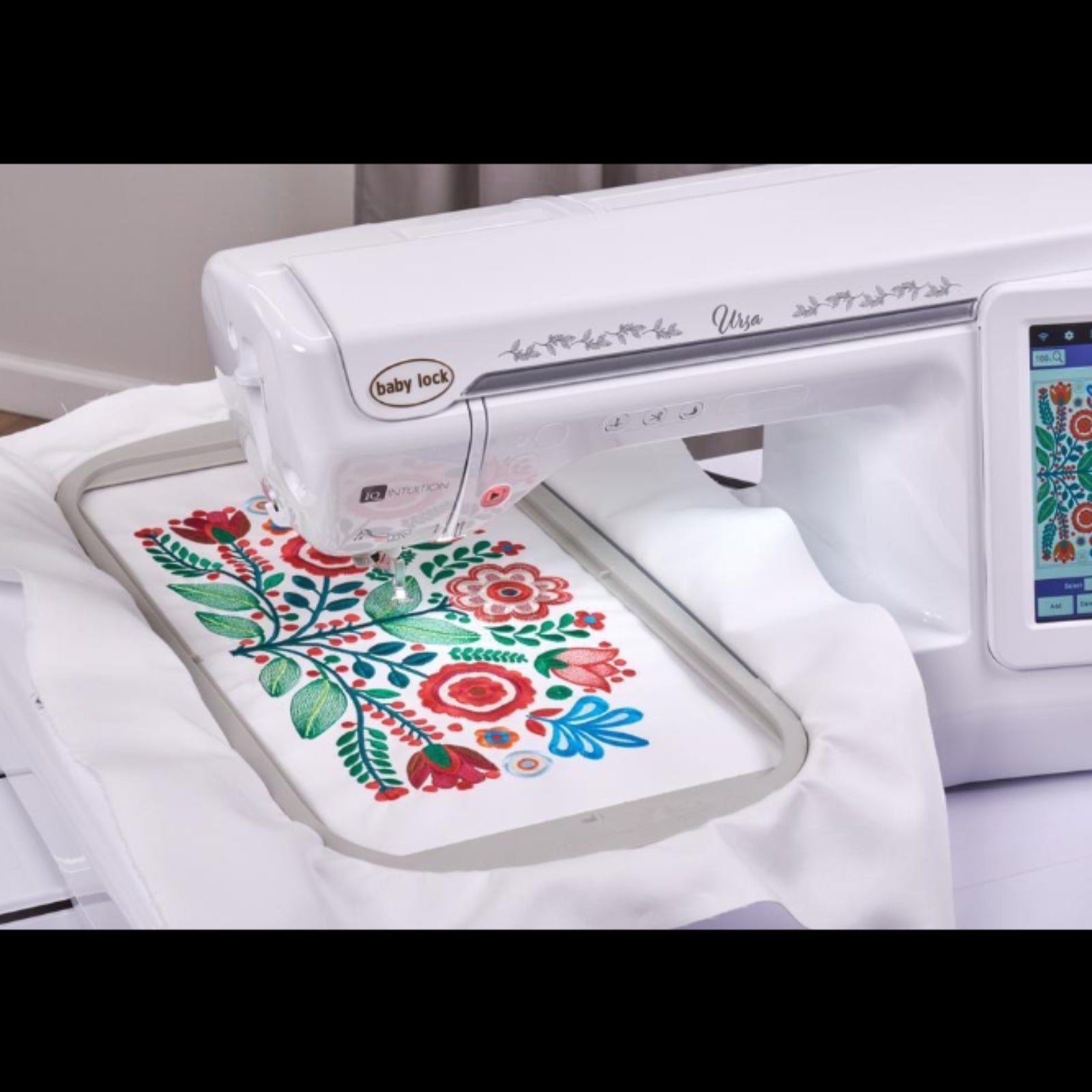 A white Baby Lock Ursa embroidery machine stitching a colorful floral design onto white fabric. The Baby Lock logo is visible on the machine head, and the screen on the right displays the embroidery pattern.