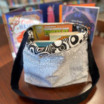 Lined book bag with a text‑print fabric design, shown filled with books on a library table.