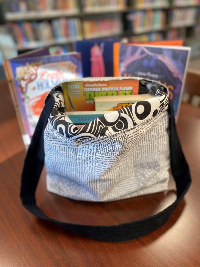 Lined book bag with a text‑print fabric design, shown filled with books on a library table.