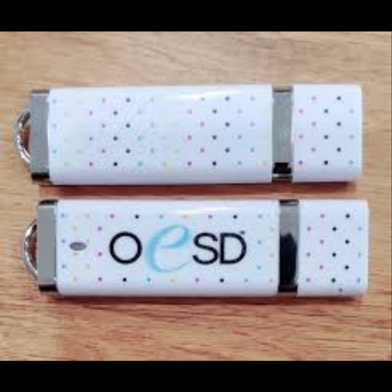 Two white USB flash drives with small multicolored polka dots and a silver metal keyring clip, placed on a light wood surface. One drive shows the text 'OeSD' in blue and gray.