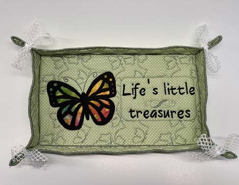Green fabric tray with lace‑trimmed corners, featuring an embroidered butterfly and the phrase “Life’s little treasures.”