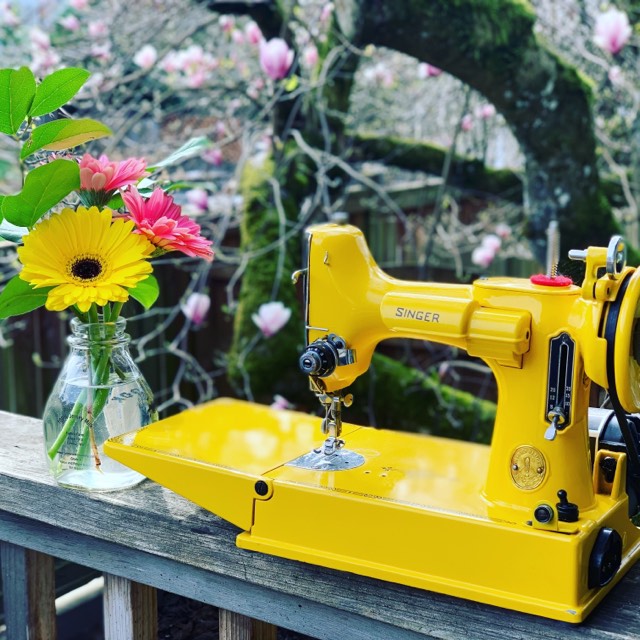 Vintage yellow Singer Featherweight set on a wooden table outdoors with a vase of bright flowers, framed by moss‑covered branches and delicate pink spring blooms.