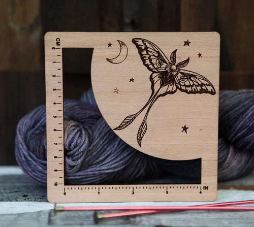 Wooden Luna Moth measuring ruler with engraved celestial details, shown with yarn and knitting needles.
