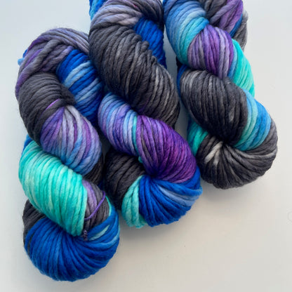 Three chunky hand‑dyed yarn skeins in deep jewel‑tone blues, purples, and grays.