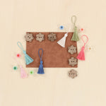 A flat-lay of small decorative craft pieces arranged on a light wood background, including several brown flower-shaped buttons, four tassels in white, light blue, blue, and pink, and several small round beads.