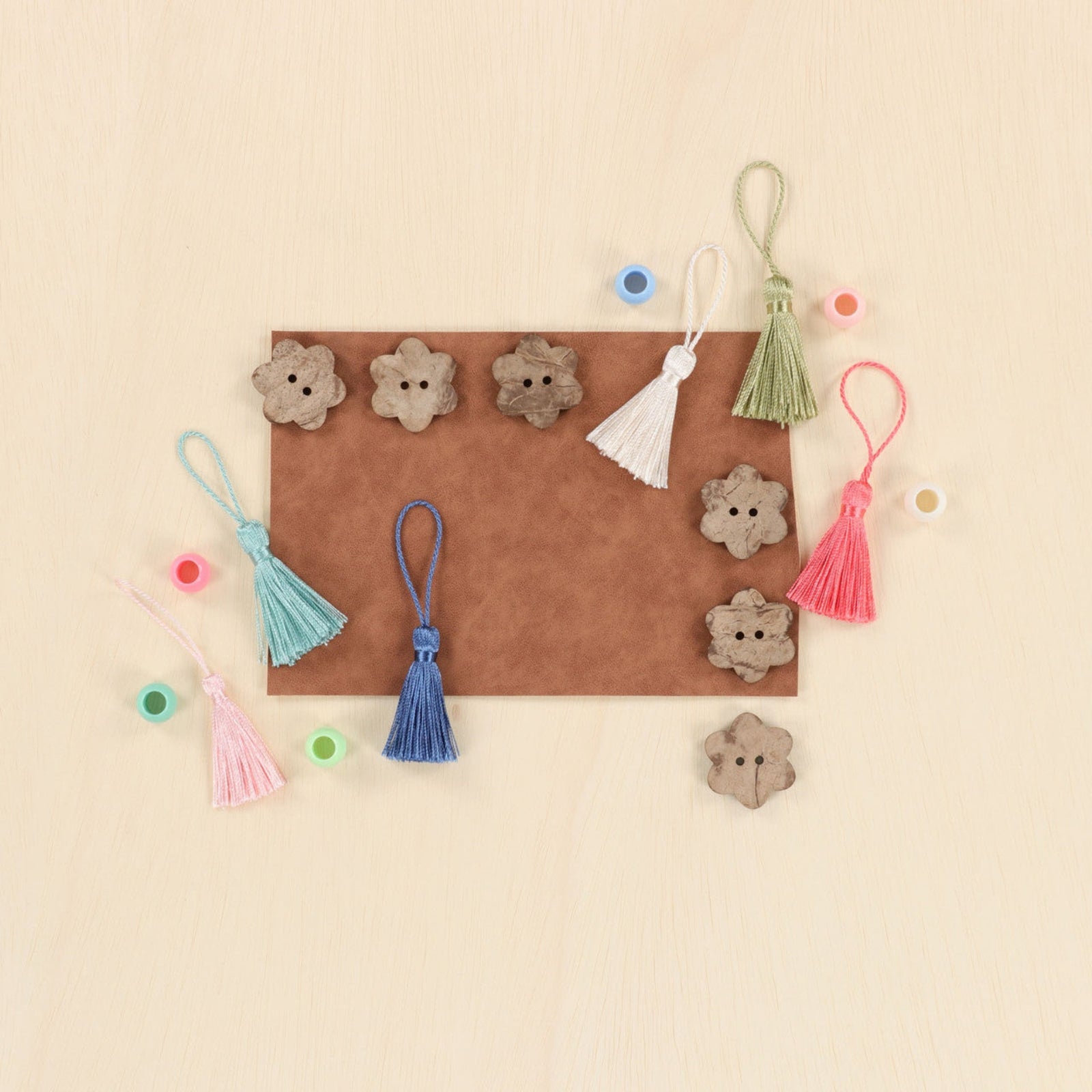 A flat-lay of small decorative craft pieces arranged on a light wood background, including several brown flower-shaped buttons, four tassels in white, light blue, blue, and pink, and several small round beads.