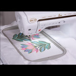 A white Baby Lock Luna embroidery machine stitching a colorful floral design inside a circular embroidery hoop on white fabric.