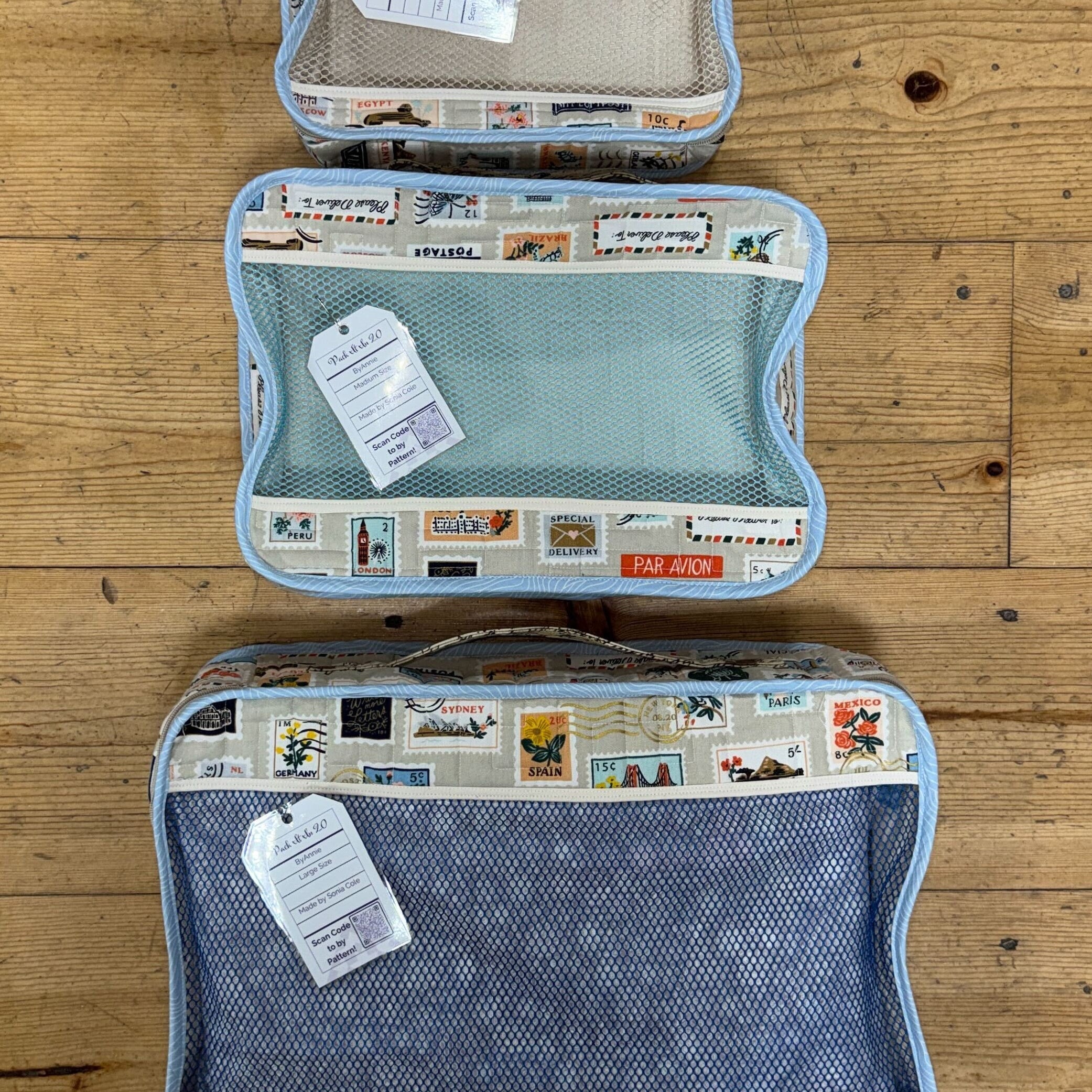 Three mesh packing cubes in small, medium, and large sizes, each with a travel‑themed postage‑stamp print border, arranged on a wooden surface.