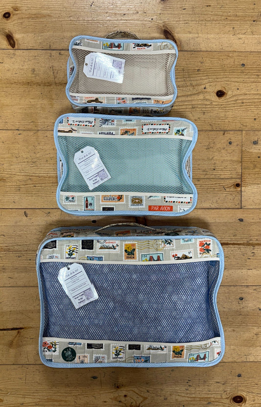 Three mesh packing cubes in small, medium, and large sizes, each with a travel‑themed postage‑stamp print border, arranged on a wooden surface.