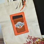 A cream-colored canvas tote bag featuring a large applique patch of a brown coffee cup with an orange polka-dot border and a white label that reads 'YOU'RE WELCOME TO Pumpkin Spice!'.
