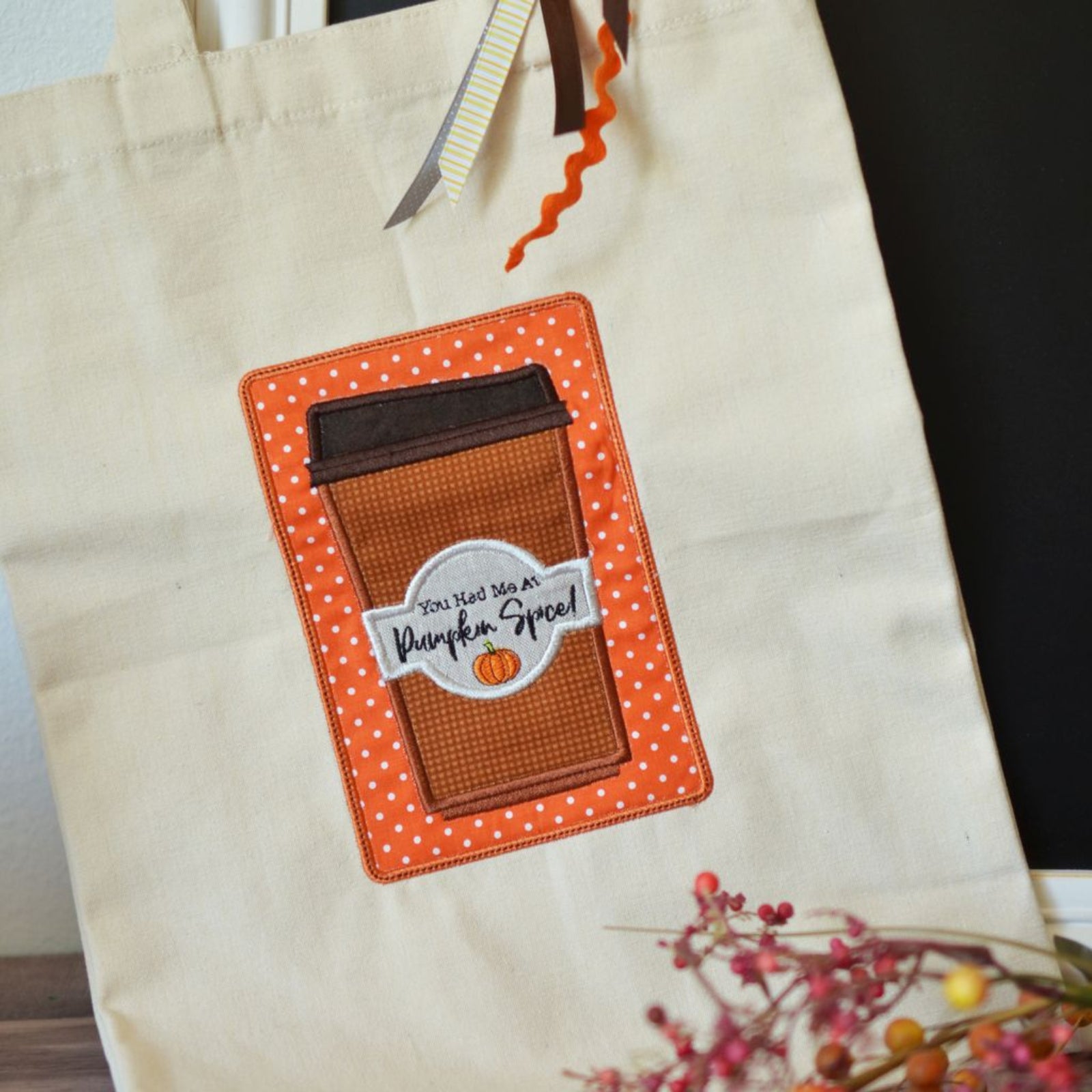 A cream-colored canvas tote bag featuring a large applique patch of a brown coffee cup with an orange polka-dot border and a white label that reads 'YOU'RE WELCOME TO Pumpkin Spice!'.