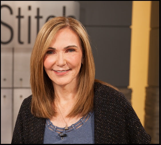 Headshot of Peggy Sagers wearing a blue top and dark cardigan, with the Fit‑2‑Stitch logo blurred in the background.