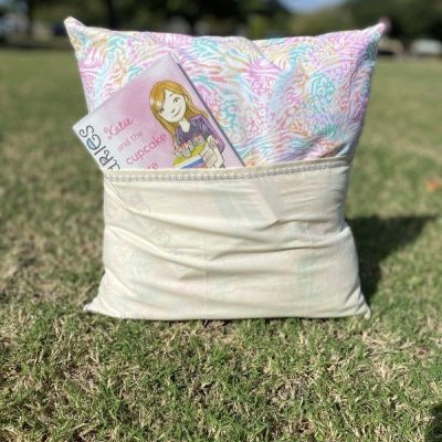 Reading pillow with a front pocket holding a children’s book, shown outdoors on the grass.