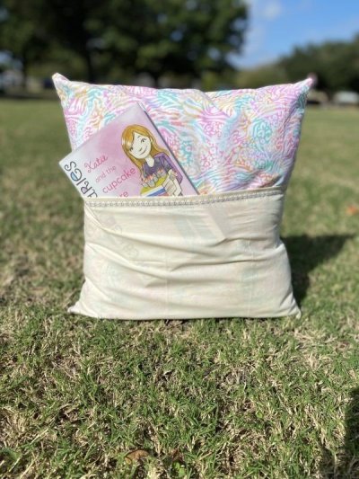 Reading pillow with a front pocket holding a children’s book, shown outdoors on the grass.