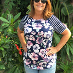 Outdoor view of the Rio Raglan with a floral front panel and striped sleeves, worn with jeans in a lush garden setting