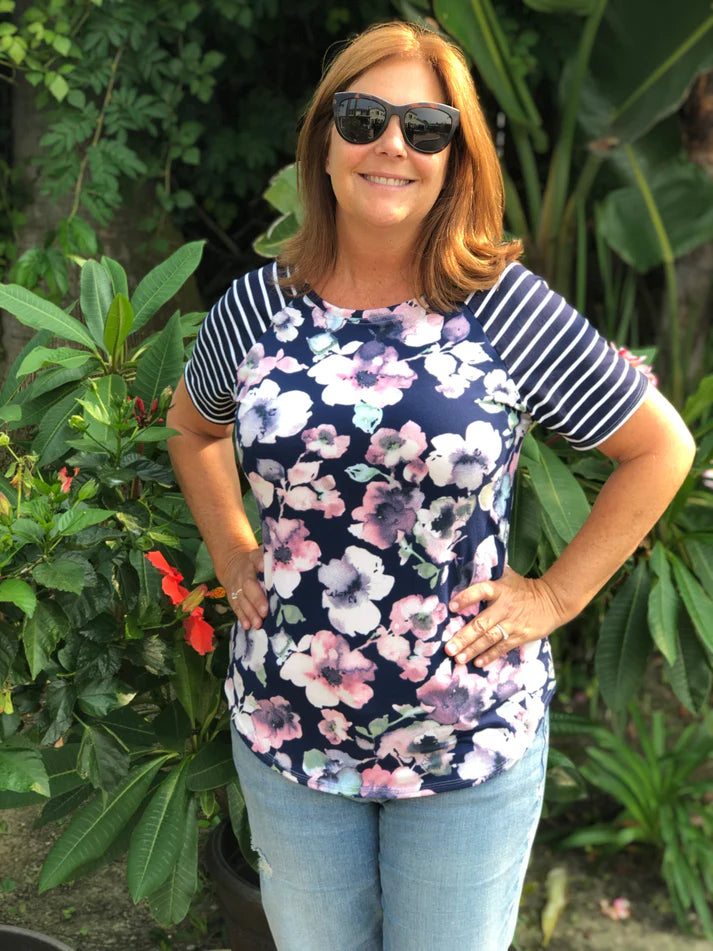 Outdoor view of the Rio Raglan with a floral front panel and striped sleeves, worn with jeans in a lush garden setting