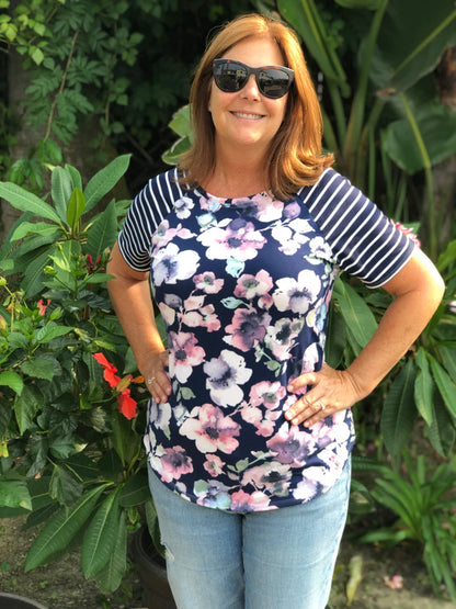 Outdoor view of the Rio Raglan with a floral front panel and striped sleeves, worn with jeans in a lush garden setting