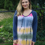Outdoor view of the Rio Raglan sewn in a dress length with long solid sleeves and a colorful tie‑dye body, shown against a garden fence backdrop
