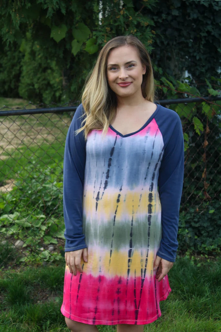 Outdoor view of the Rio Raglan sewn in a dress length with long solid sleeves and a colorful tie‑dye body, shown against a garden fence backdrop
