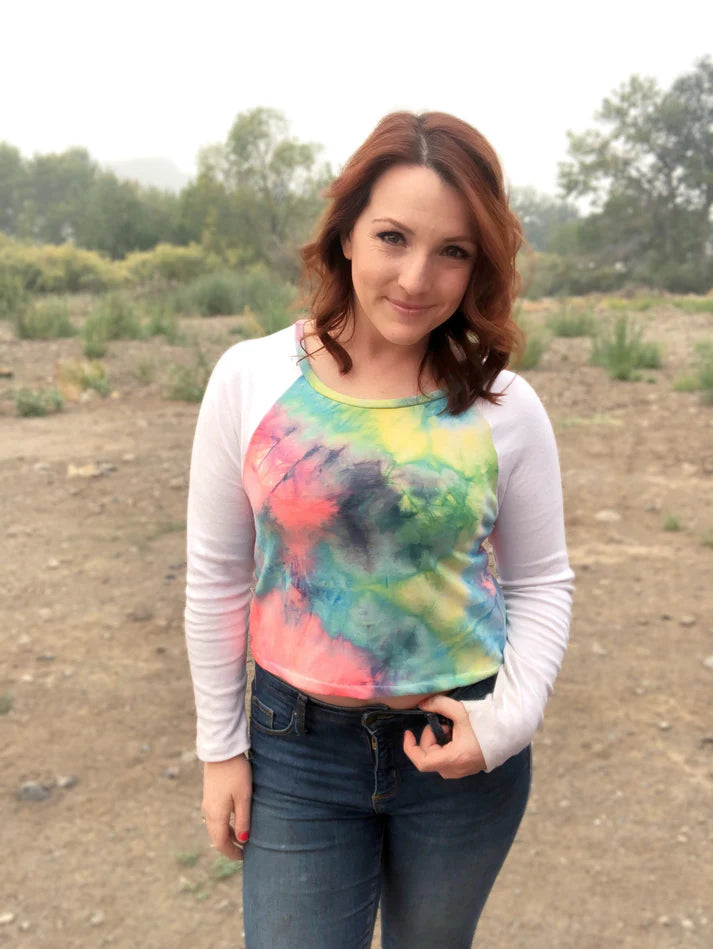 Outdoor view of the Rio Raglan in a long‑sleeve tie‑dye front with solid white sleeves, worn with jeans in a natural setting