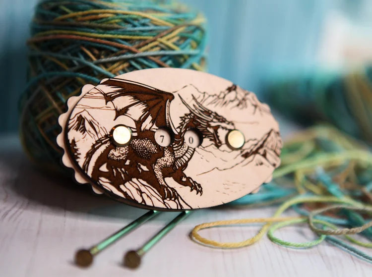 Wooden dragon‑engraved row counter with number dials, shown beside multicolored yarn and knitting needles.