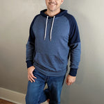 Indoor view of the Santa Fe Hoodie sewn as a pullover with a hood, featuring blue and navy color‑blocking and white drawstrings, worn with jeans against a light wall