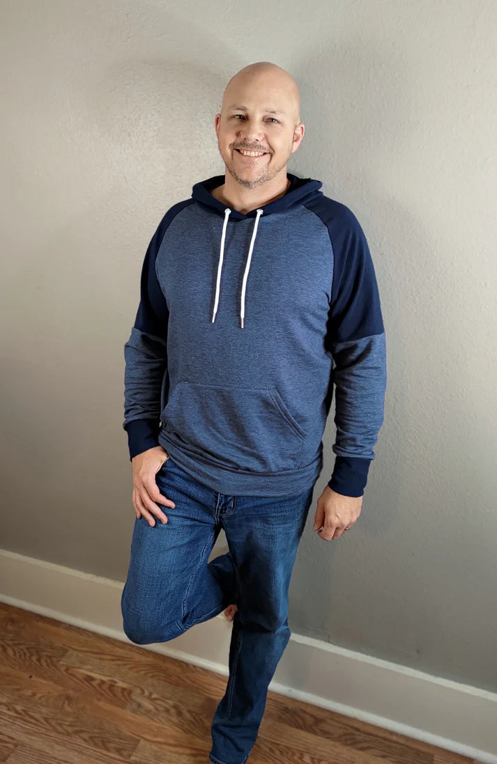 Indoor view of the Santa Fe Hoodie sewn as a pullover with a hood, featuring blue and navy color‑blocking and white drawstrings, worn with jeans against a light wall