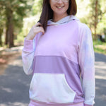 Outdoor view of the Santa Fe Hoodie sewn with long sleeves, a hood, and a pink‑to‑purple color‑block design, worn on a forested path