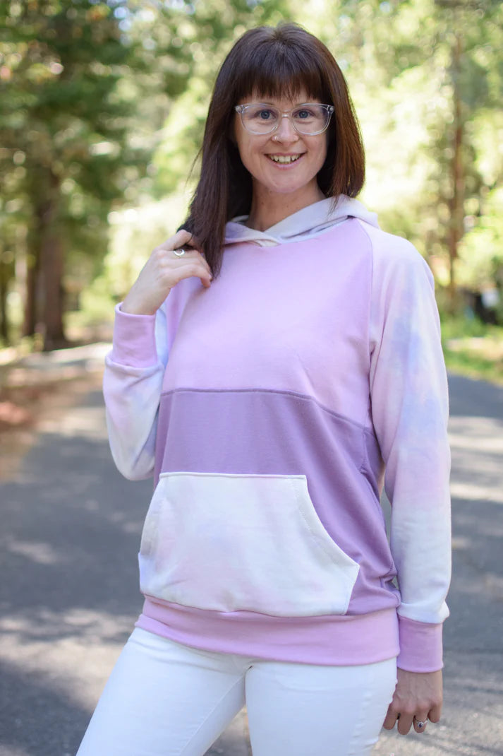 Outdoor view of the Santa Fe Hoodie sewn with long sleeves, a hood, and a pink‑to‑purple color‑block design, worn on a forested path