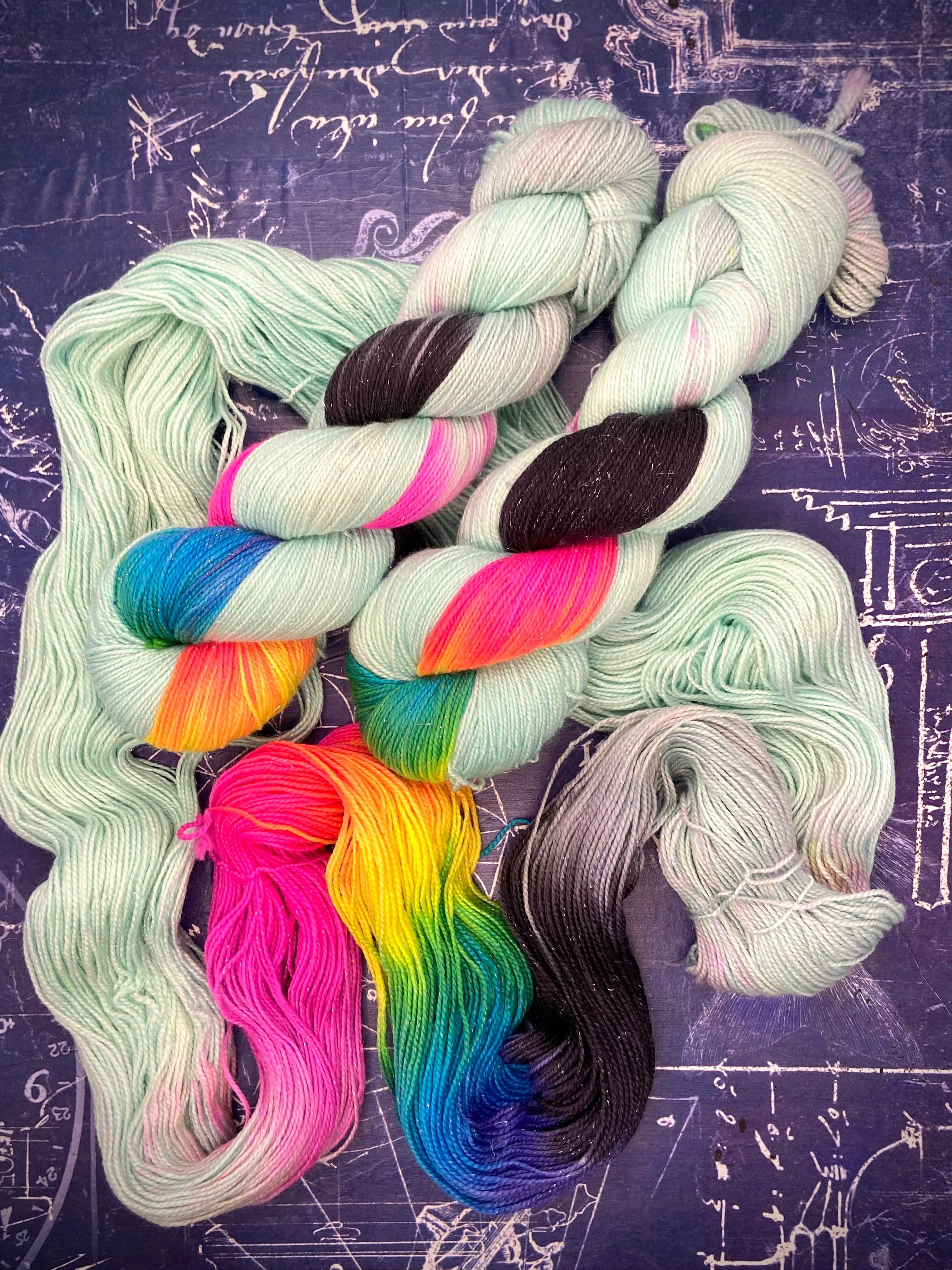 Pale mint hand‑dyed yarn with bright rainbow and black sections arranged on a dark schematic background.