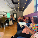 A woman knitting on a blue couch during Stitch ’n Bitch, with Rachel and other makers gathered in the background at Barley Pop Brewing.